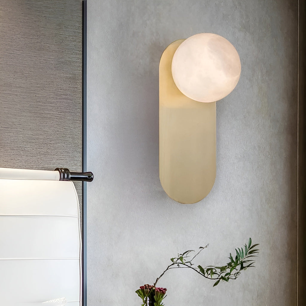 Adela wall sconce seen at an angle on a grey wall, its alabaster shade lit above a spray of greenery