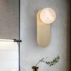 Adela wall sconce seen at an angle on a grey wall, its alabaster shade lit above a spray of greenery