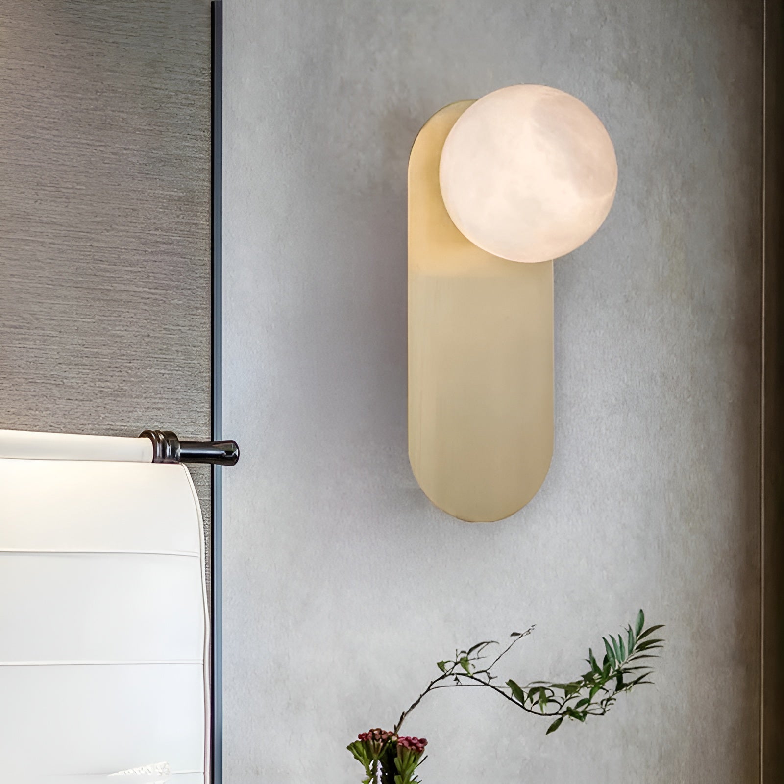 Adela wall sconce seen at an angle on a grey wall, its alabaster shade lit above a spray of greenery
