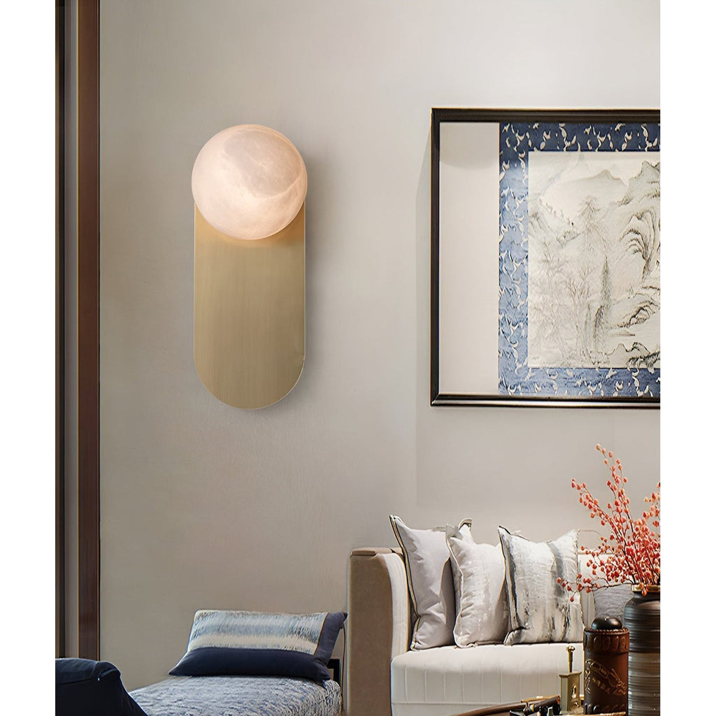 Adela wall sconce glowing warm in a living room beside a framed landscape painting and a cushioned sofa