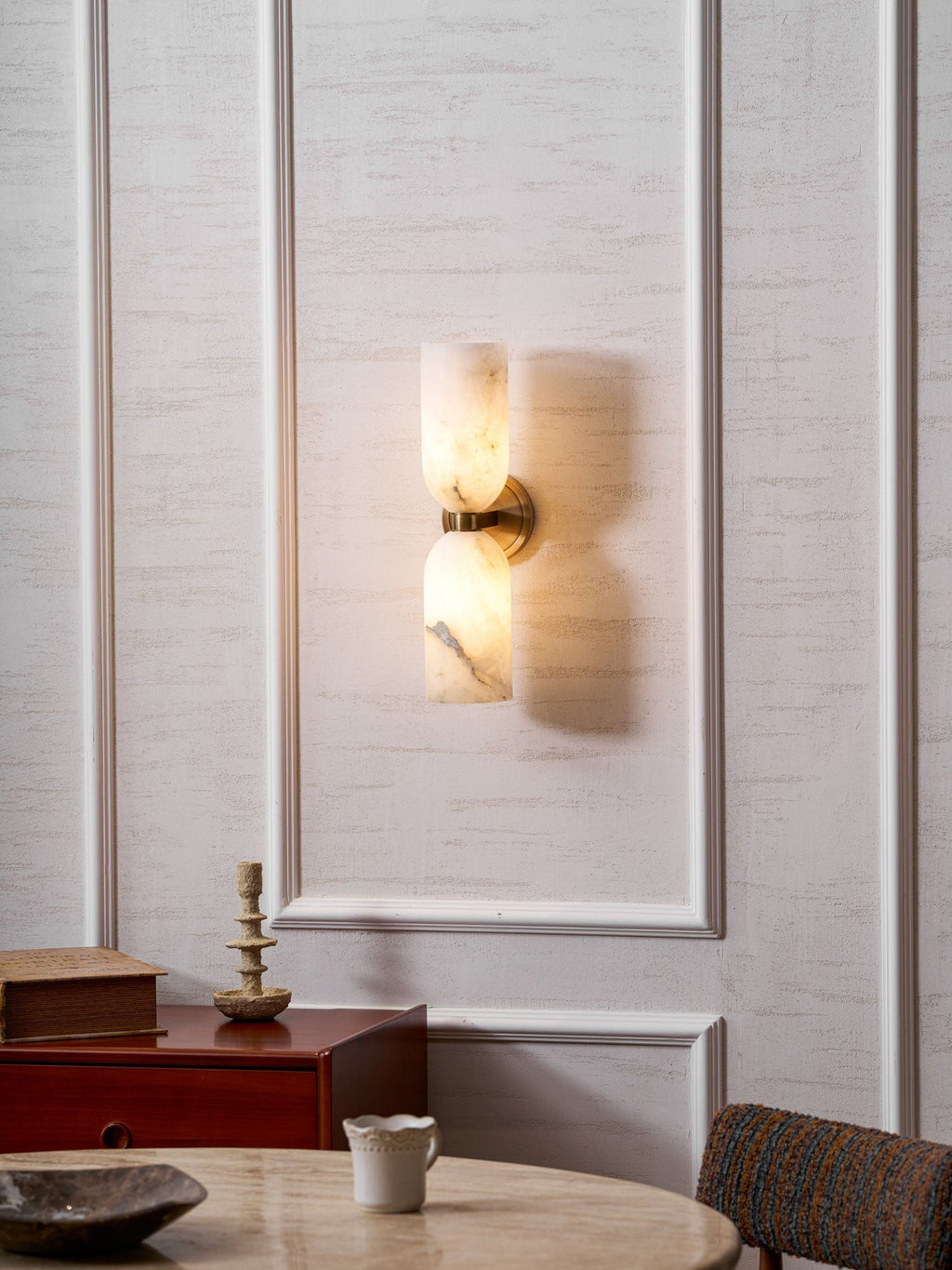 The lit Nexus Modern Alabaster Wall Light on a panelled wall beside a wooden sideboard and dining table.