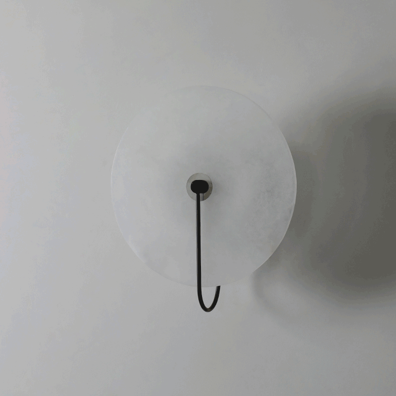 The Kristy wall lamp unlit on a plain wall, black arm hooking below the pale alabaster disc.
