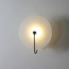 The Elaris wall light in black, its slim hook arm crossing the glowing alabaster disc face.