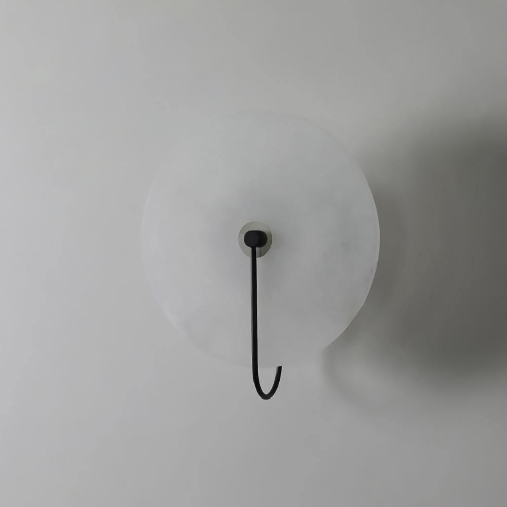 Unlit black Elaris wall light on a white wall, the hook arm curving down past the disc edge.