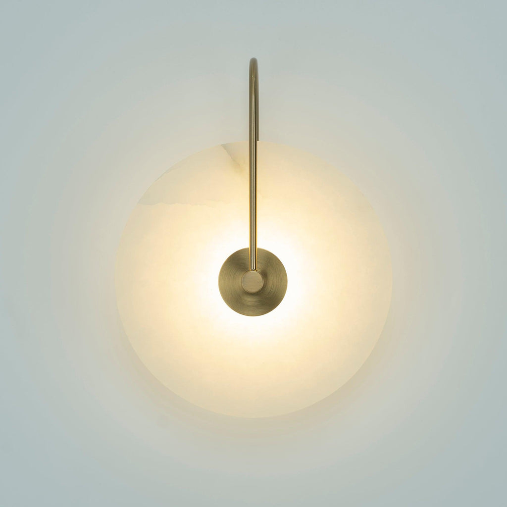 Elaris wall light with an antique brass arm, lit and glowing warm on a pale blue-grey wall.