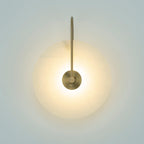 Elaris wall light with an antique brass arm, lit and glowing warm on a pale blue-grey wall.