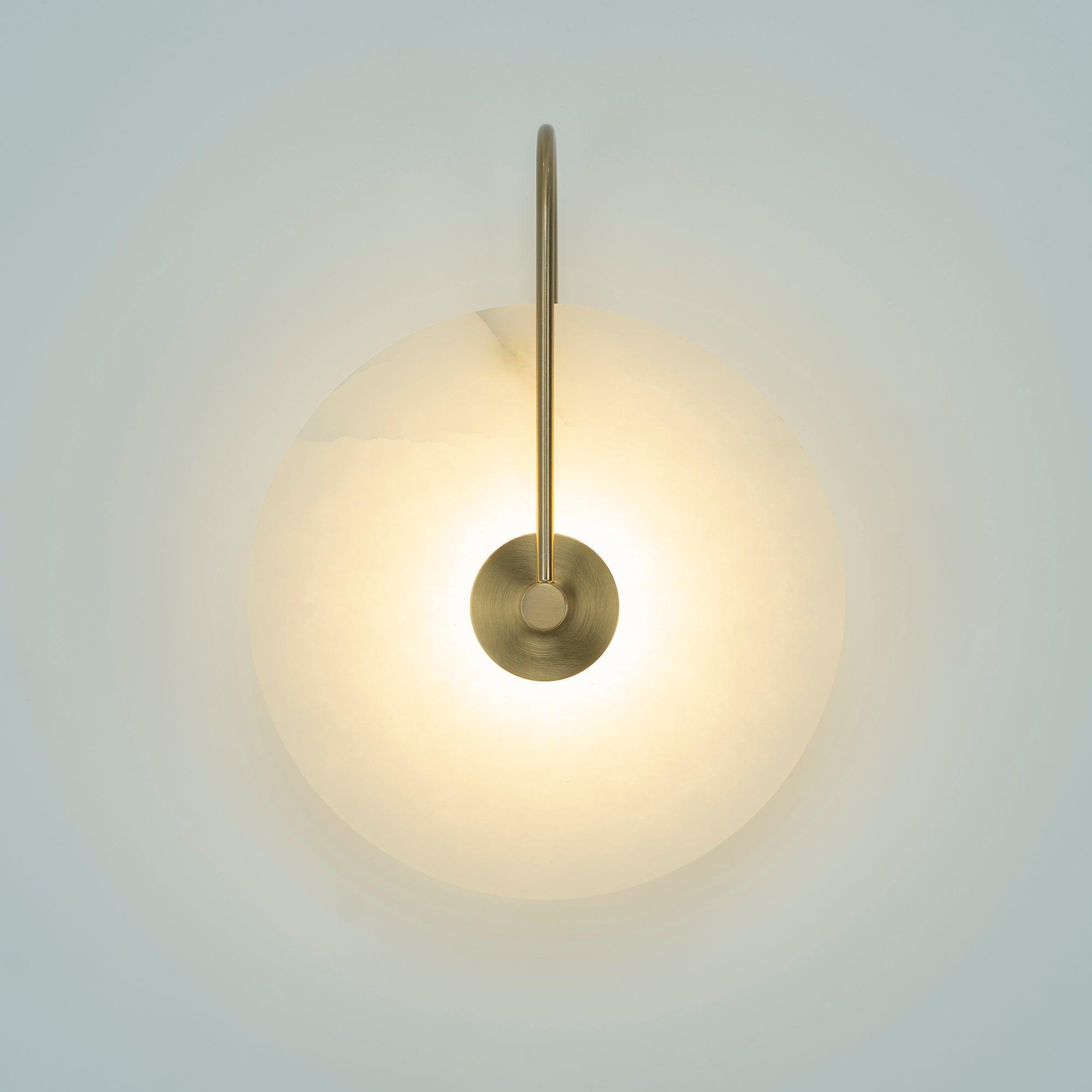 Elaris wall light with an antique brass arm, lit and glowing warm on a pale blue-grey wall.