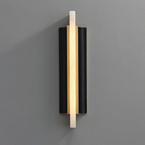 Aurelian alabaster wall sconce lit on a grey wall, its stone blade glowing between brass and black panels
