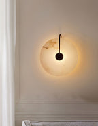 Black Elaris wall light lit on a panelled cream wall beside a curtain, its glow ringing the disc.