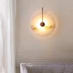 Gold Elaris wall light lit on a panelled cream wall, casting a wide halo above a grey sofa.