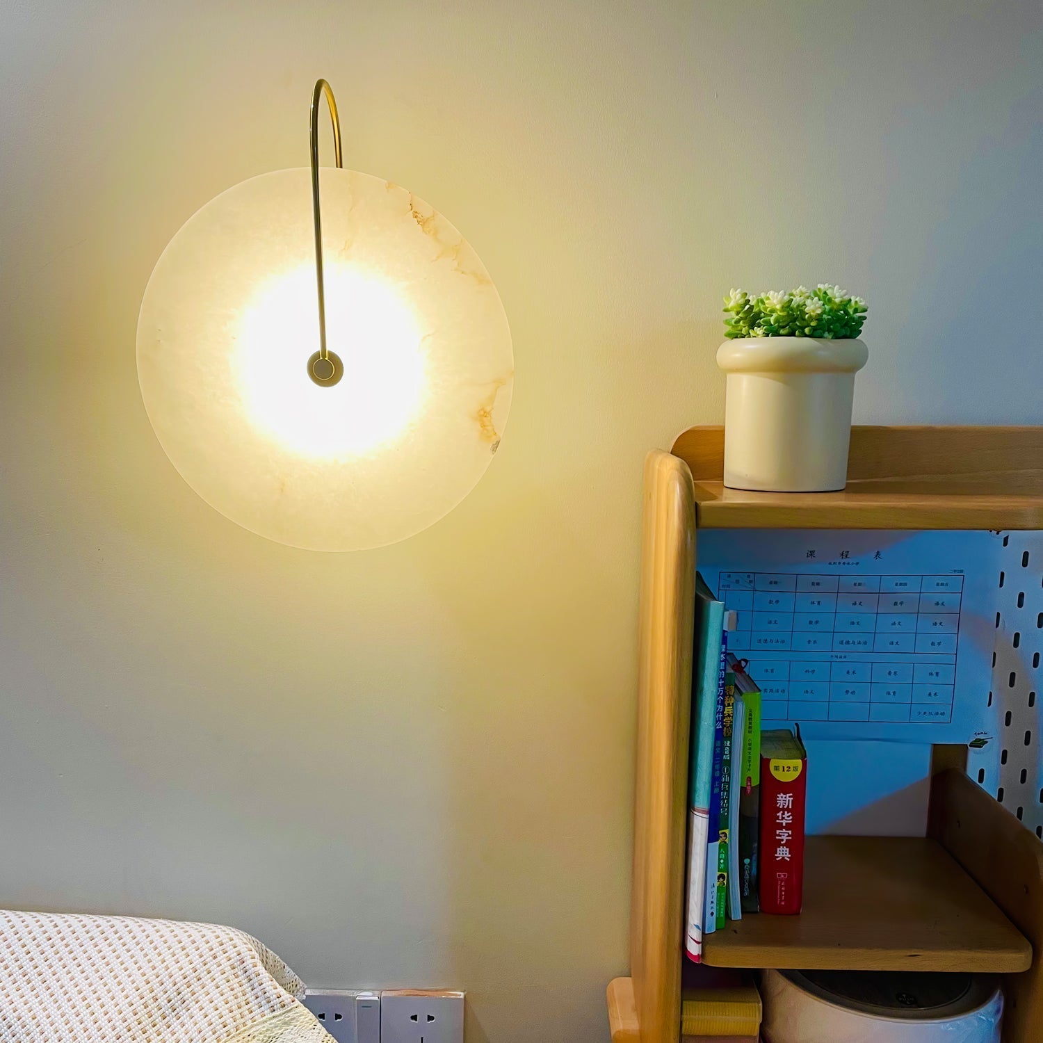 Gold Elaris wall light lit in a real bedroom beside a wooden bookshelf and a small potted plant.