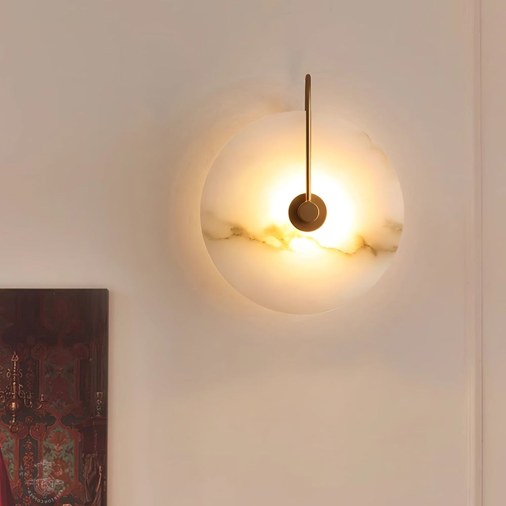 Kristy wall lamp lit on a pale wall beside a framed painting, veining glowing across the alabaster disc.