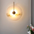 Gold Elaris wall light lit on a cream wall near a corner moulding, with lilies in the foreground.