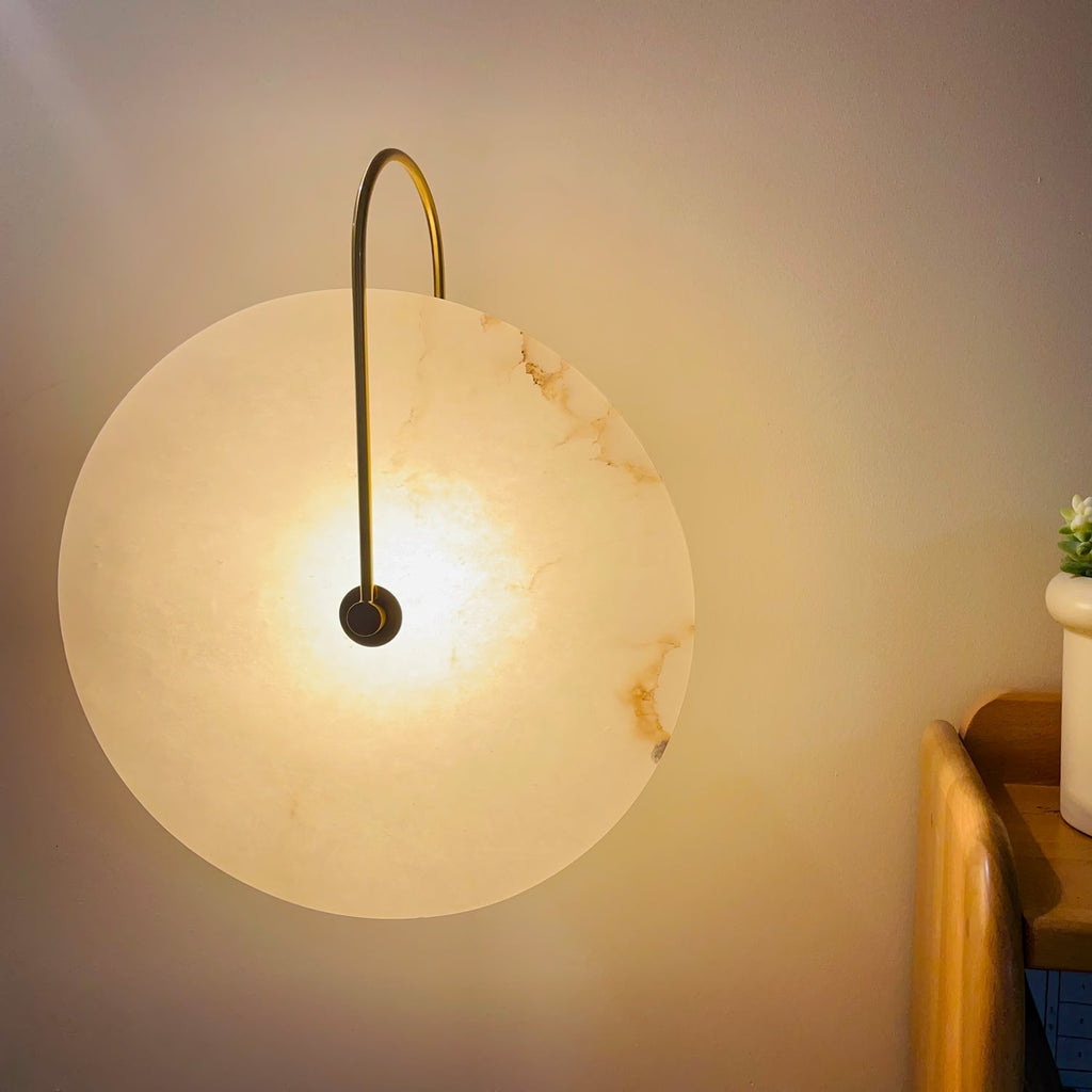 Gold Elaris wall light lit in a real bedroom corner, warm light washing the wall by a bedside shelf.