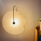 Gold Elaris wall light lit in a real bedroom corner, warm light washing the wall by a bedside shelf.
