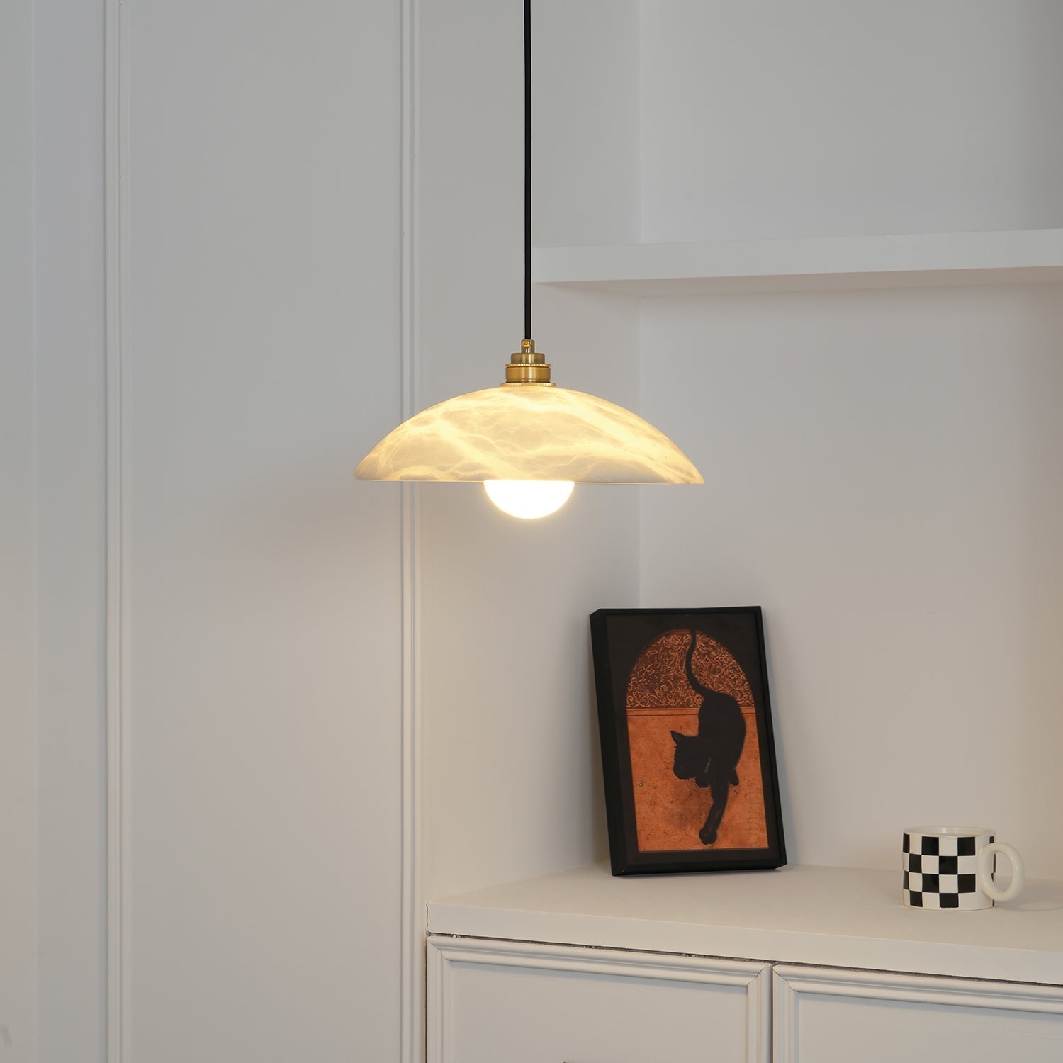 Warm-lit Ritta alabaster dome pendant above a white cabinet with a framed cat print and mug.