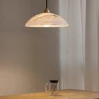 Nerea pendant lit above a wooden table with a glass mug, sheer curtain drawn behind it.