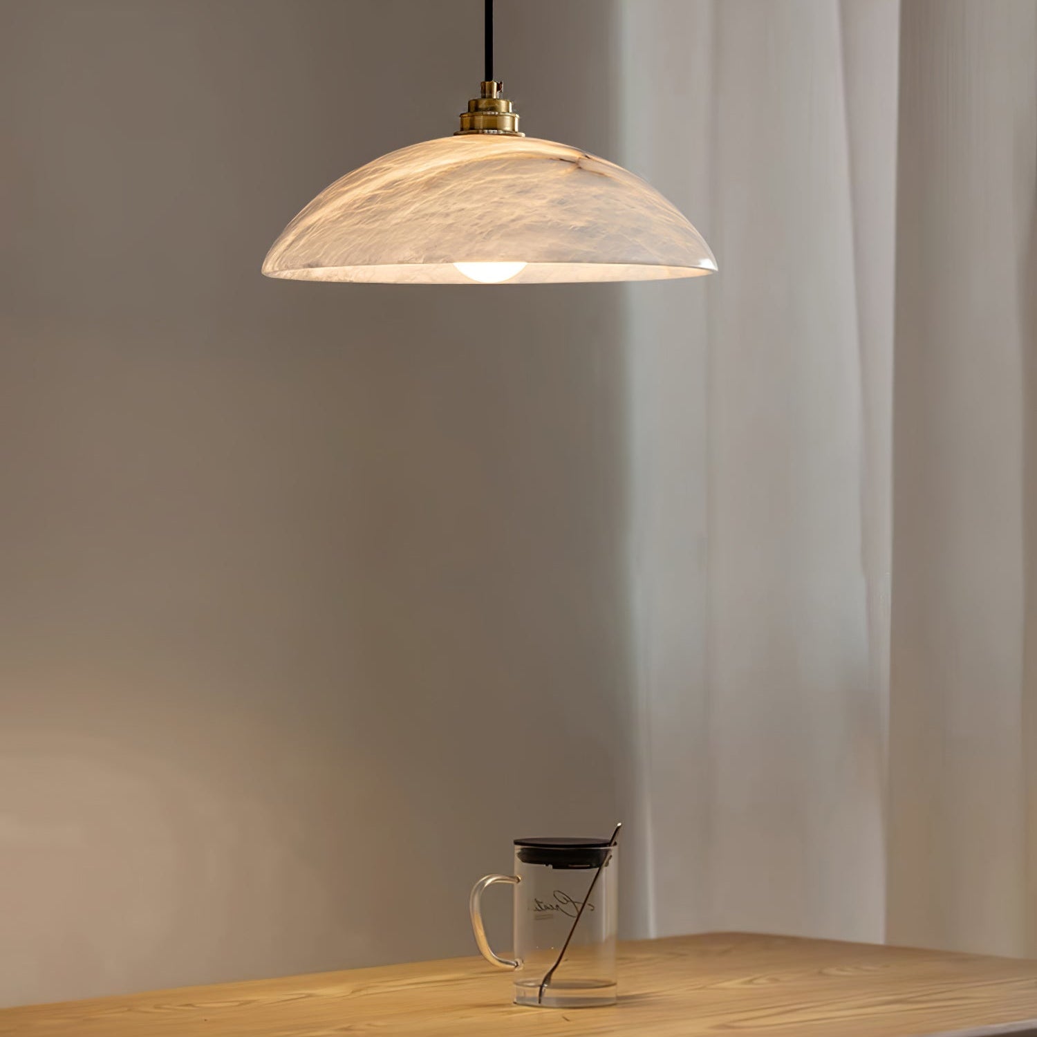 Nerea pendant lit above a wooden table with a glass mug, sheer curtain drawn behind it.