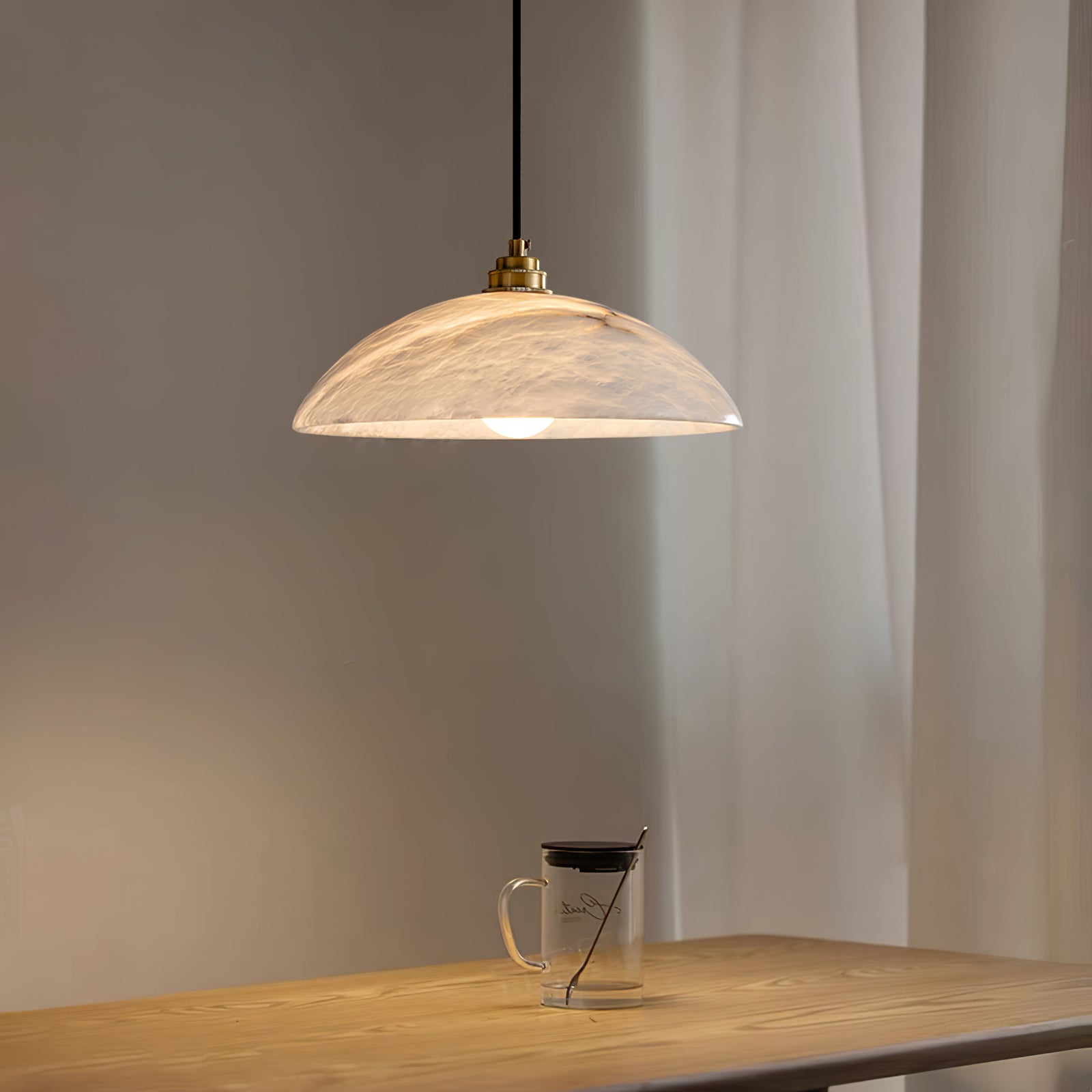 Ritta alabaster dome pendant lighting a wooden table with a glass mug beside a sheer curtain.