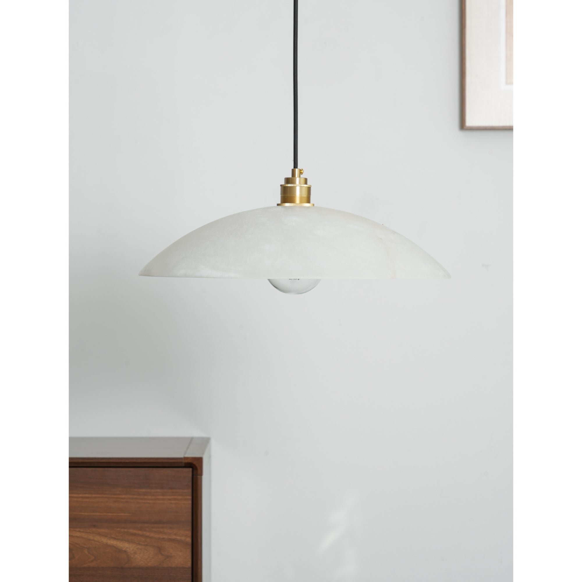 Nerea pendant unlit against a white wall, walnut chest and framed artwork behind it.