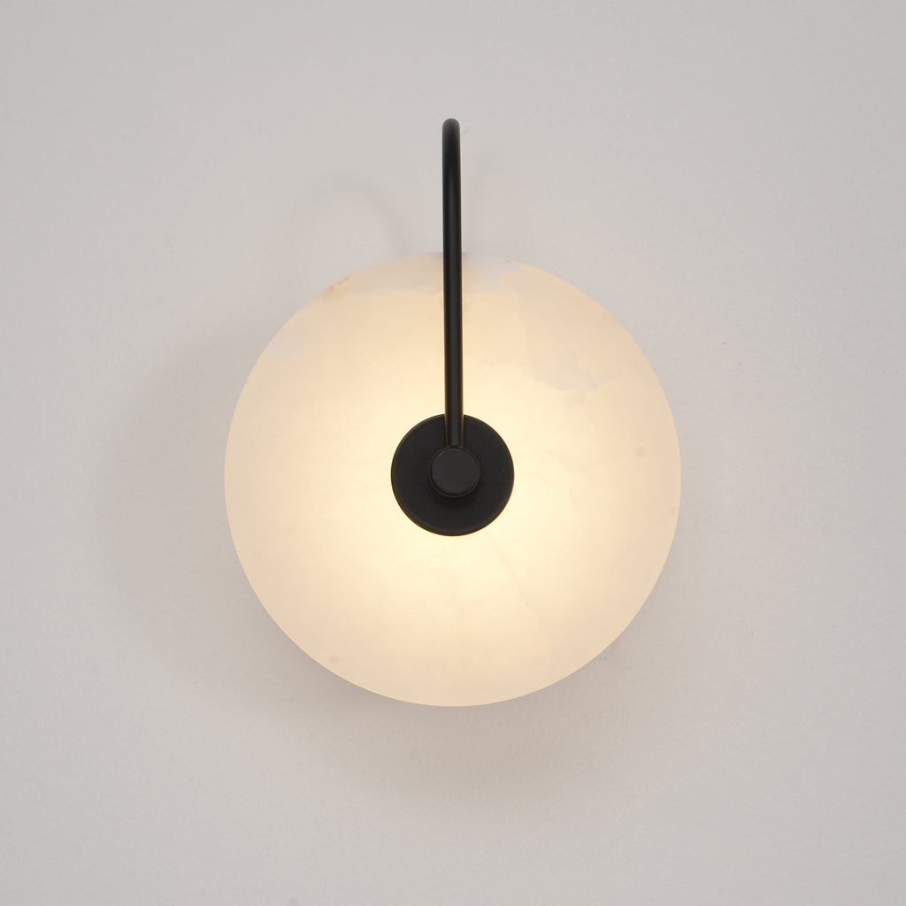 Black-arm Elaris wall light lit front-on, its warm glow spreading evenly across the alabaster disc.