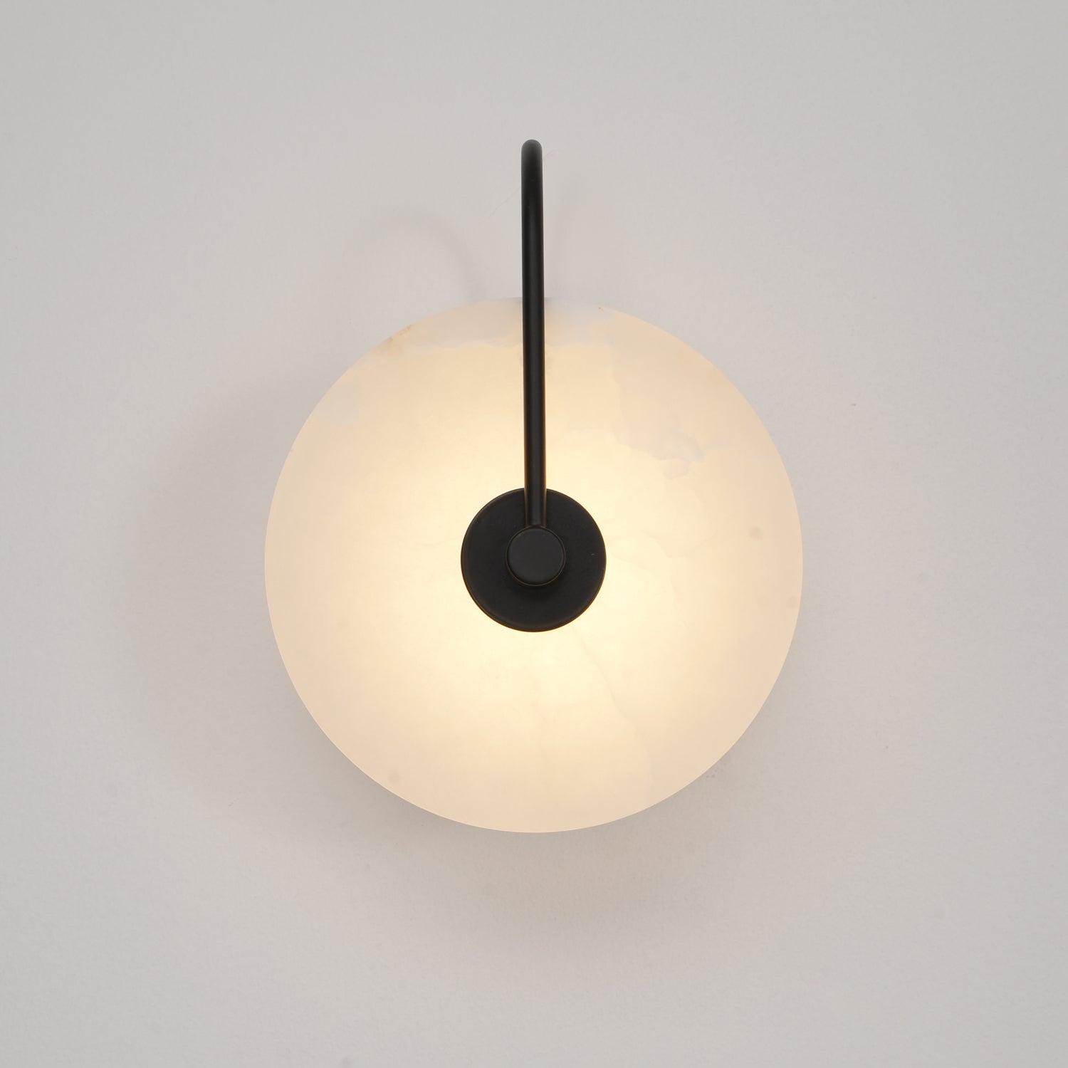 Black-arm Elaris wall light lit front-on, its warm glow spreading evenly across the alabaster disc.