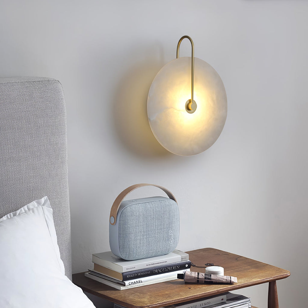Gold-arm Elaris wall light lit above a wooden bedside table with books, next to a grey headboard.