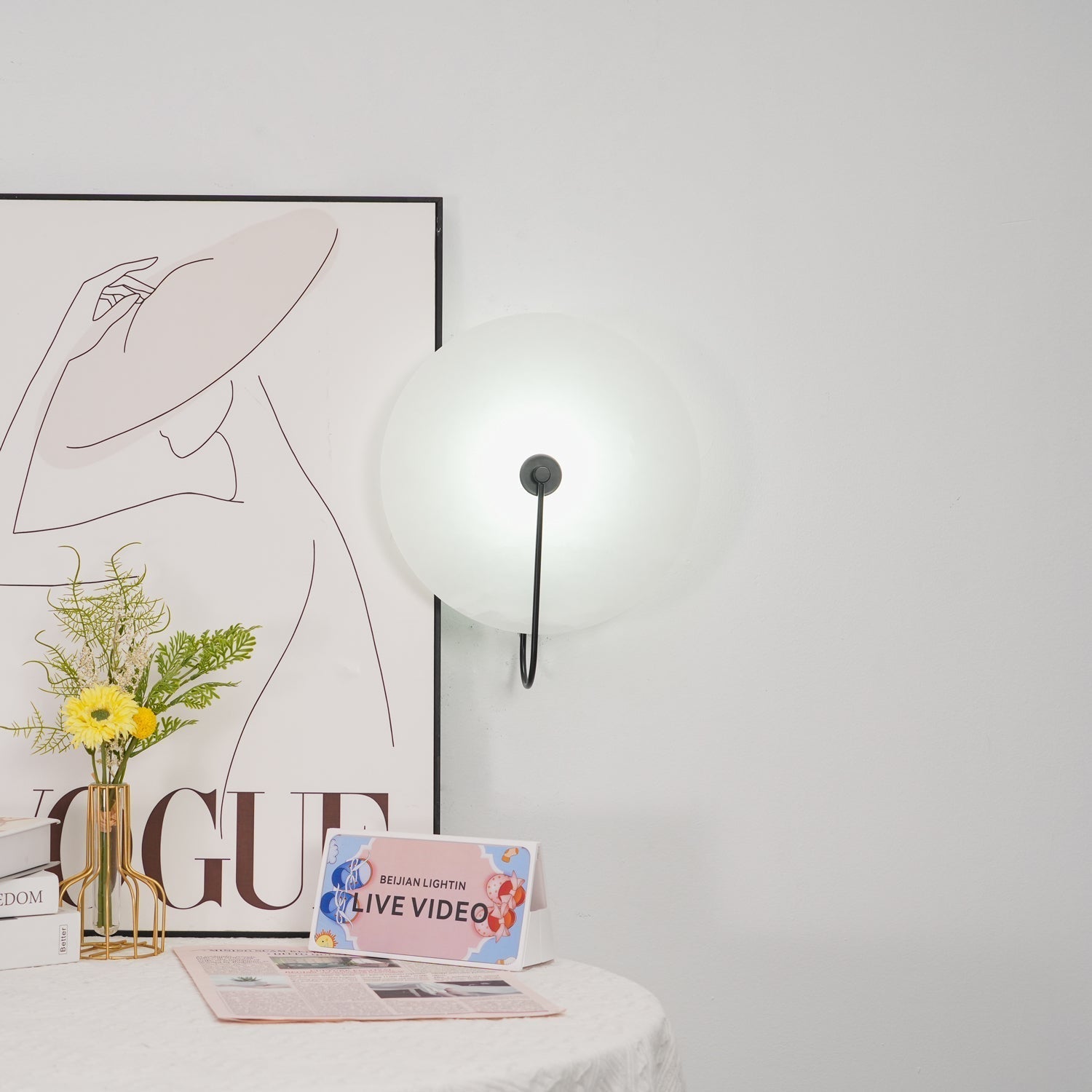 Wider view of the black Elaris wall light unlit on a white wall next to a framed print and vase.