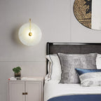 Elaris wall light in gold lit above a bedside table, beside a black bed with a rattan headboard.