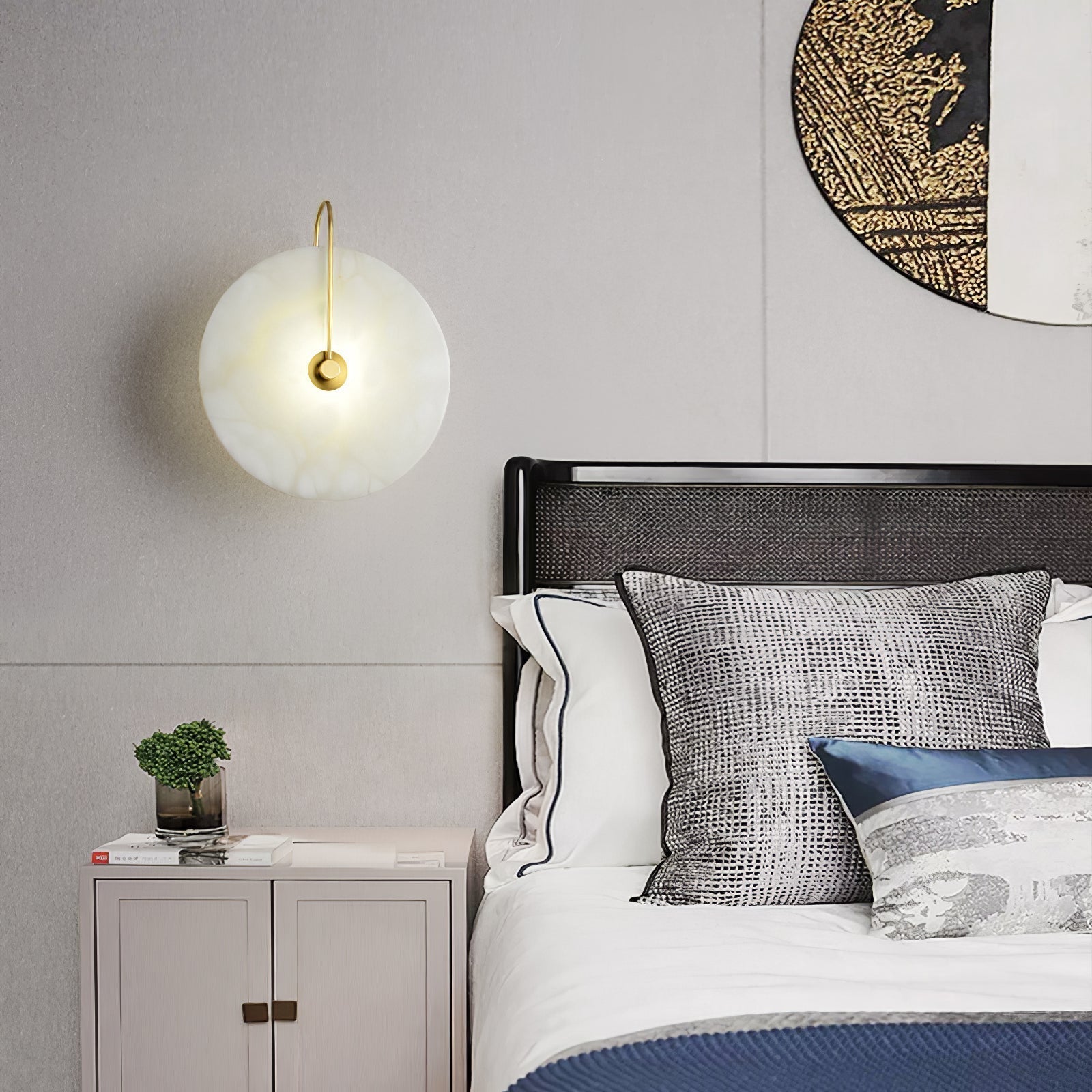 Elaris wall light in gold lit above a bedside table, beside a black bed with a rattan headboard.