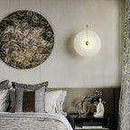 Gold Elaris wall light lit beside a large round artwork above a layered, cushion-piled bed.