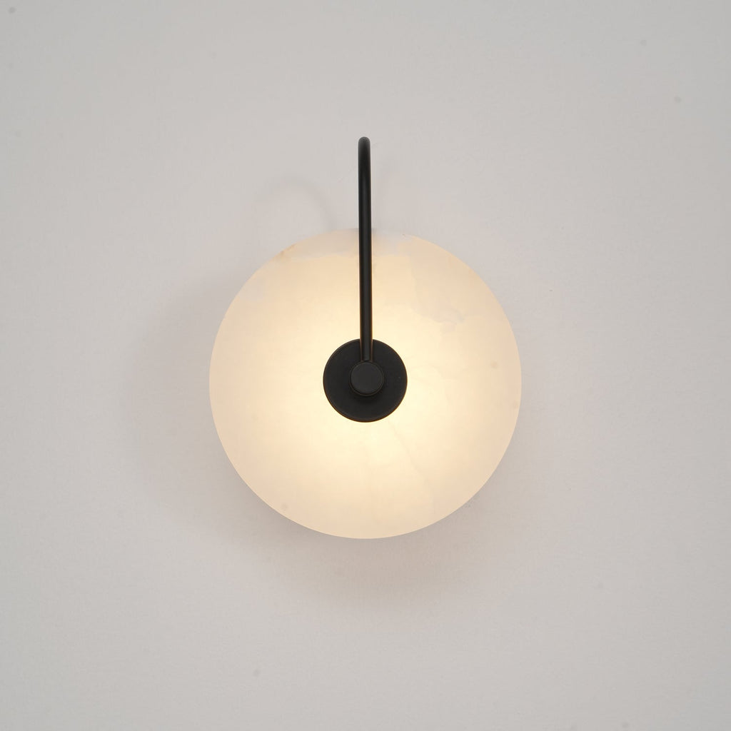 Black Elaris wall light lit low and soft, the disc face evenly bright against an off-white wall.