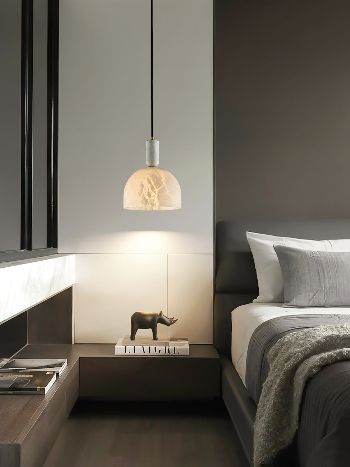 Clérachic alabaster pendant light lit above a built-in wooden nightstand holding a small book and ornament, beside taupe bedding.