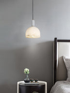 Clérachic alabaster pendant light beside a dark tufted headboard, hanging over a round side table with a small vase of flowers.