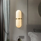Clérachic alabaster wall lamp lit beside a bed, brass band catching the light next to a grey headboard