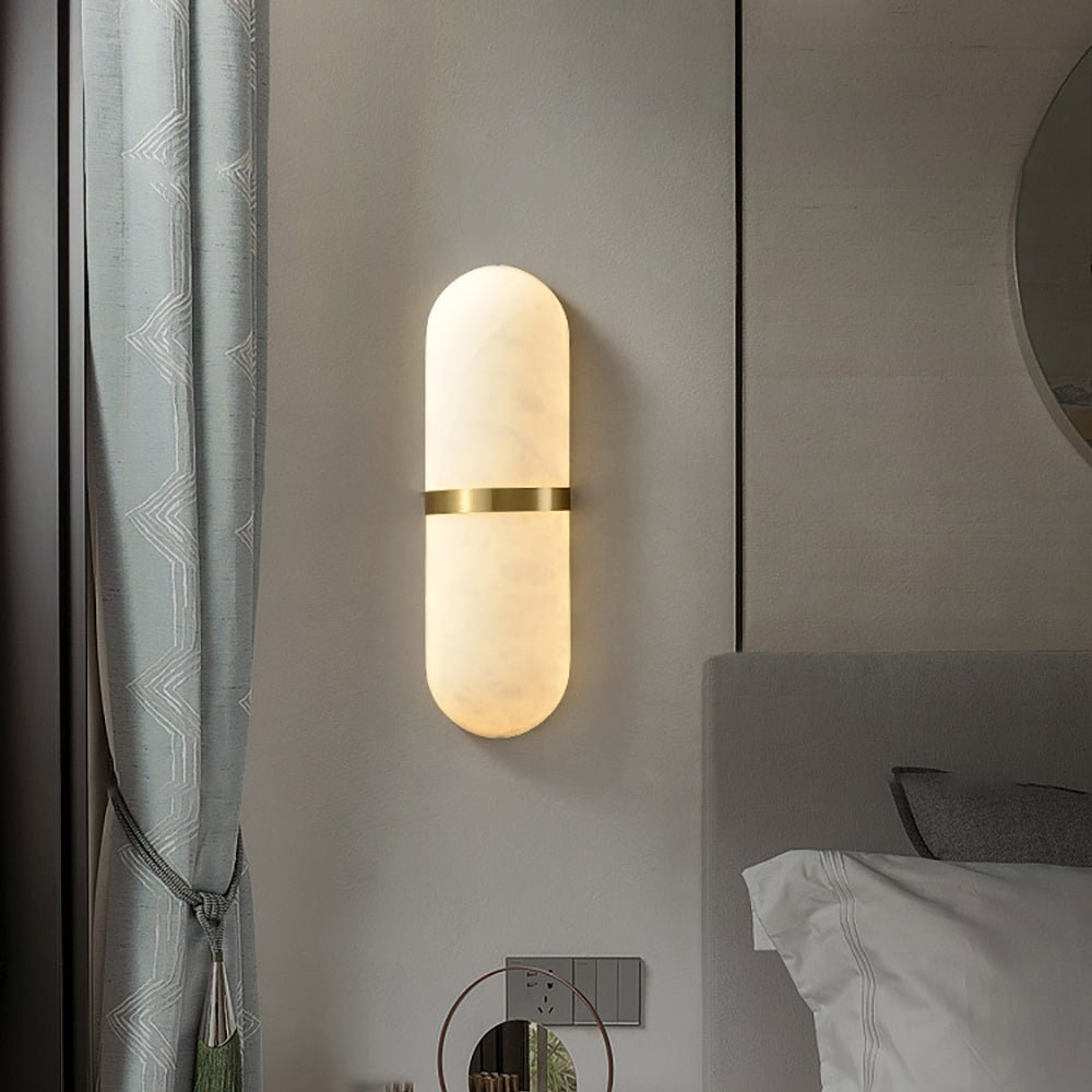 Clérachic alabaster wall lamp lit beside a bed, brass band catching the light next to a grey headboard
