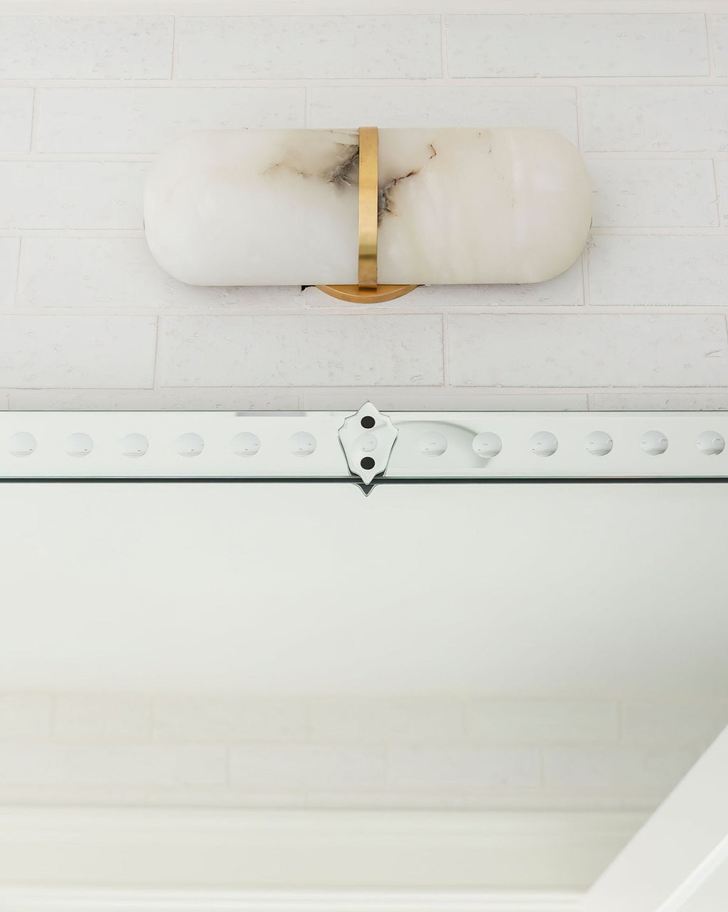 The Clérachic alabaster wall lamp mounted horizontally above a mirror, brass band centred, on white brick