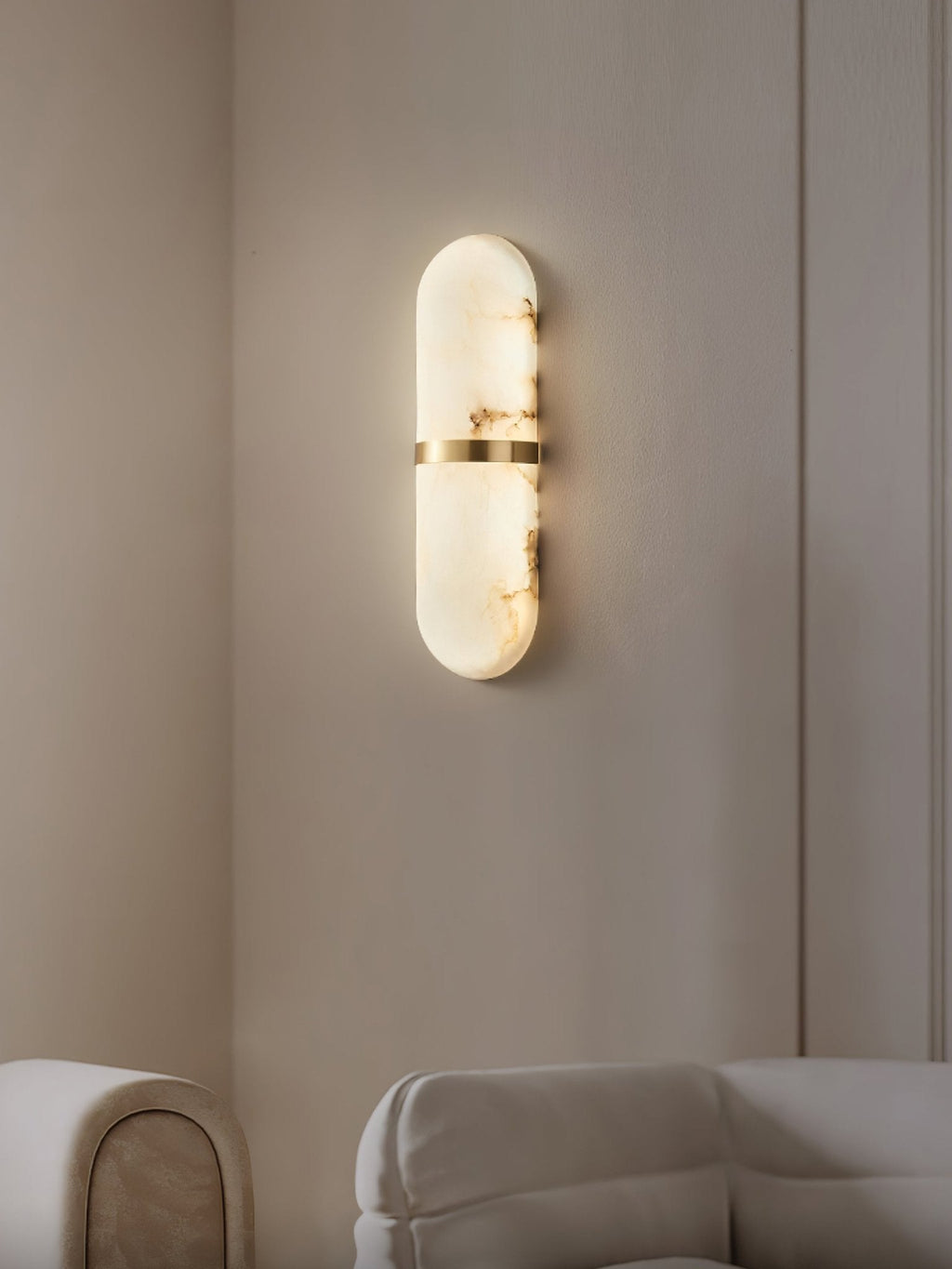 Clérachic alabaster wall lamp lit in a beige panelled corner above a cream sofa, warm glow on the wall
