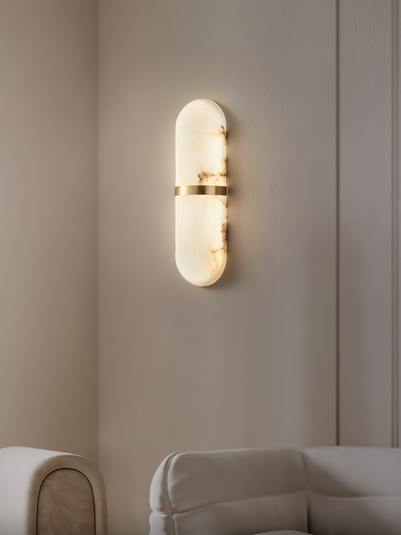 Clérachic alabaster wall lamp lit in a beige panelled corner above a cream sofa, warm glow on the wall