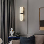 Clérachic black-banded alabaster wall lamp lit on a grey panelled living-room wall beside velvet curtains