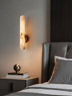 The sconce lit against a grey wall next to a bed, brass ball ornament on books below.
