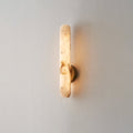 Alabaster rod sconce lit on a pale cream wall, its brass disc mount centred on the stone bar.