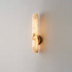 Alabaster rod sconce lit on a pale cream wall, its brass disc mount centred on the stone bar.