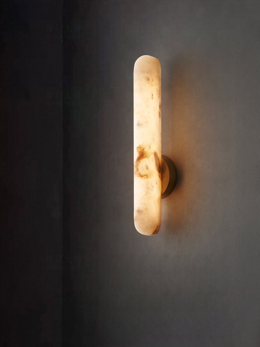The lit rod sconce on a deep grey wall, its veining glowing amber through the alabaster.