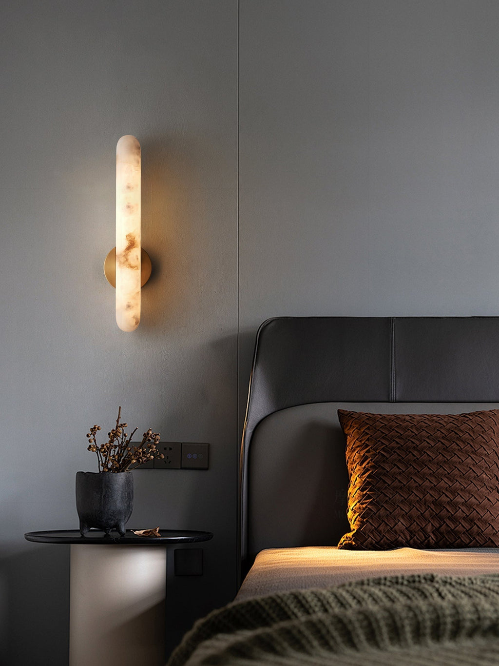 Lit beside a leather headboard, a black round table with dried flowers below the sconce.