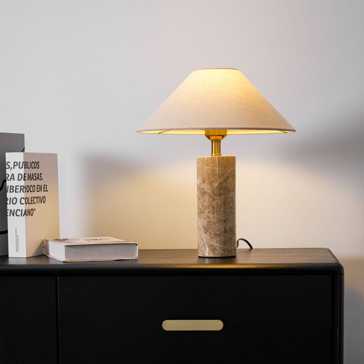 The Clérachic table lamp lit on a black dresser beside a stack of books and a black leather chair edge in the room's corner.
