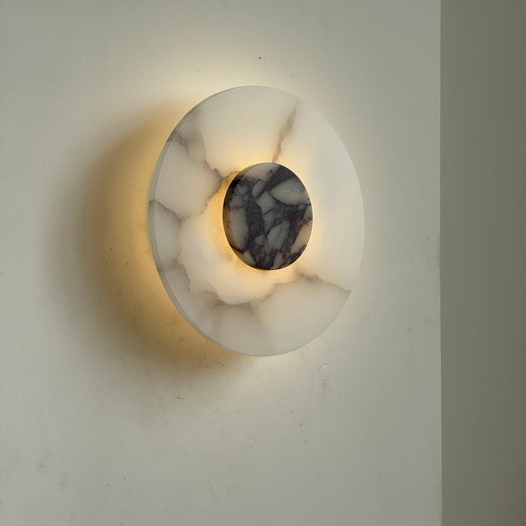 Clérachic rauduvyi wall lamp seen at an angle, lit, with light washing the wall behind the alabaster disc