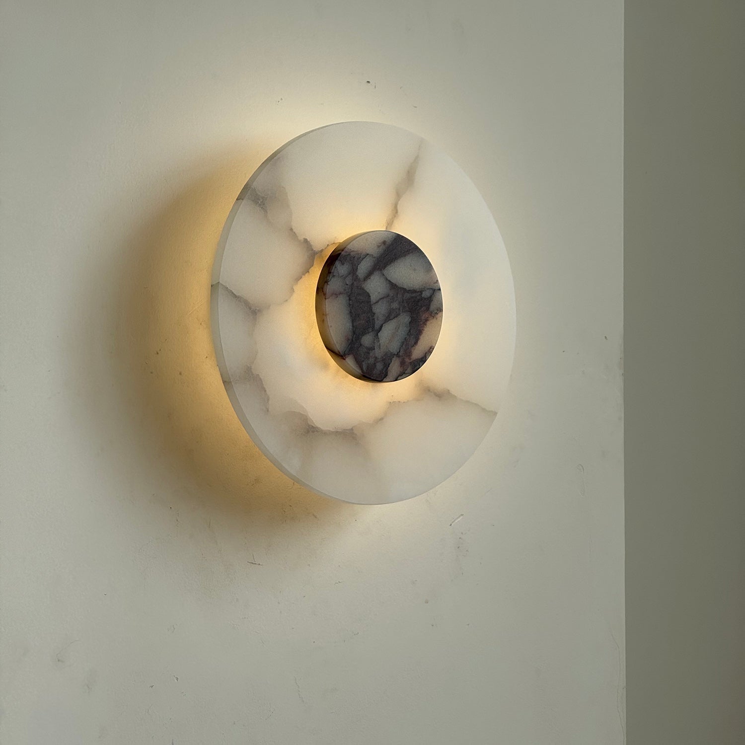 Clérachic rauduvyi wall lamp seen at an angle, lit, with light washing the wall behind the alabaster disc