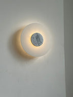 Clérachic rauduvyi wall lamp lit at an angle, warm light spilling around the grey-veined white marble disc