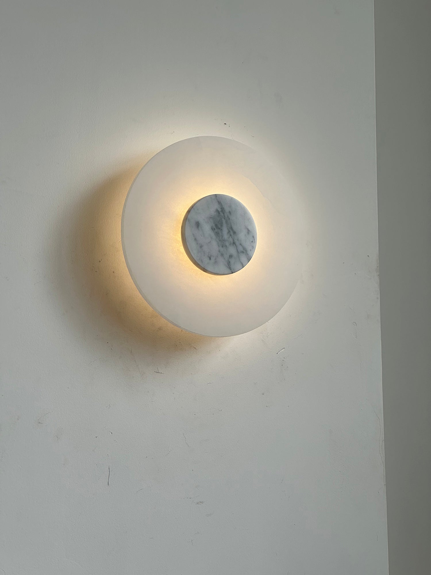 Clérachic rauduvyi wall lamp lit at an angle, warm light spilling around the grey-veined white marble disc